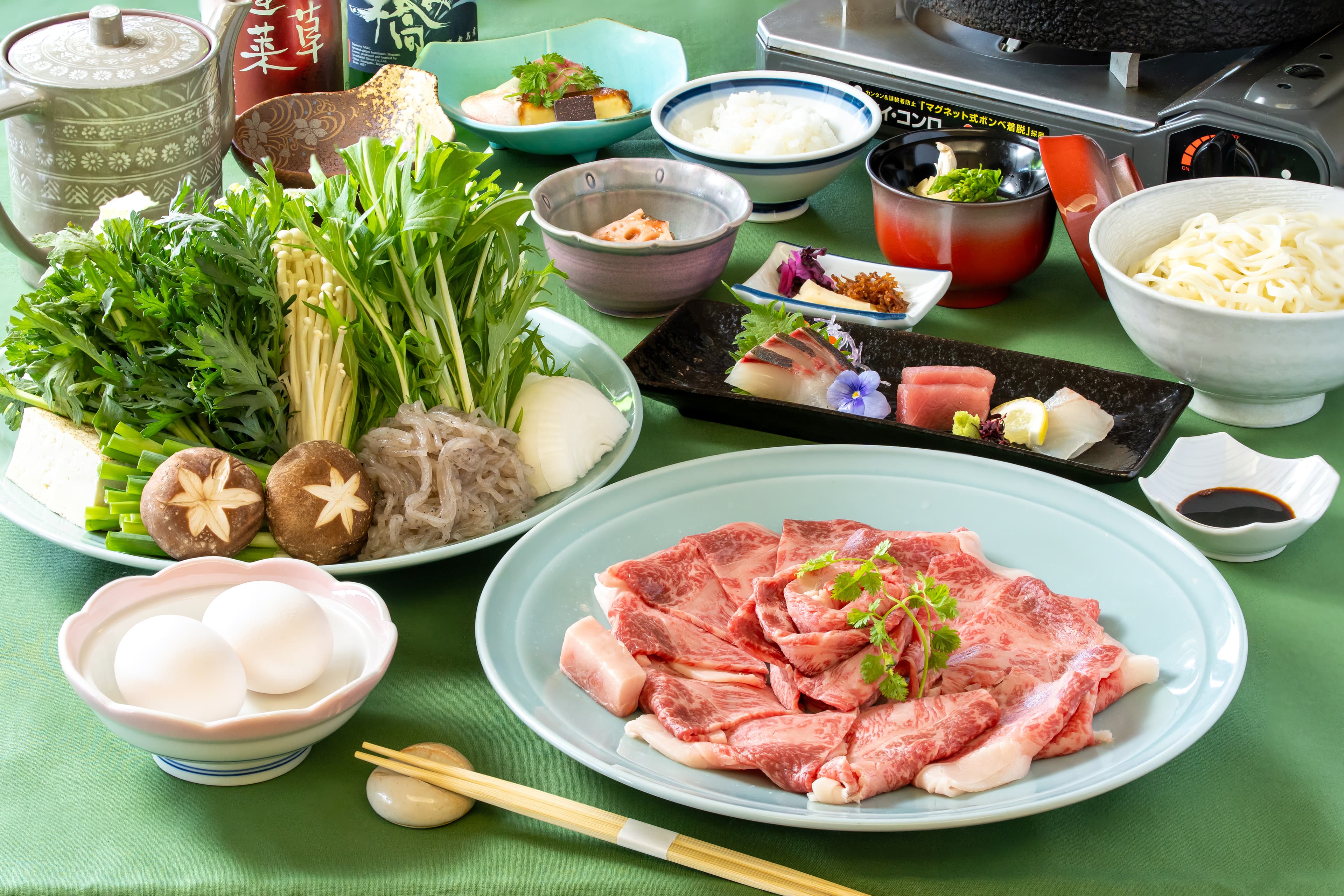 強羅荘のすき焼きコースの膳。和牛サーロインのスライスと鍋野菜、造り、うどんやご飯、卵、調理用の鍋とコンロがテーブルに並んだ様子。